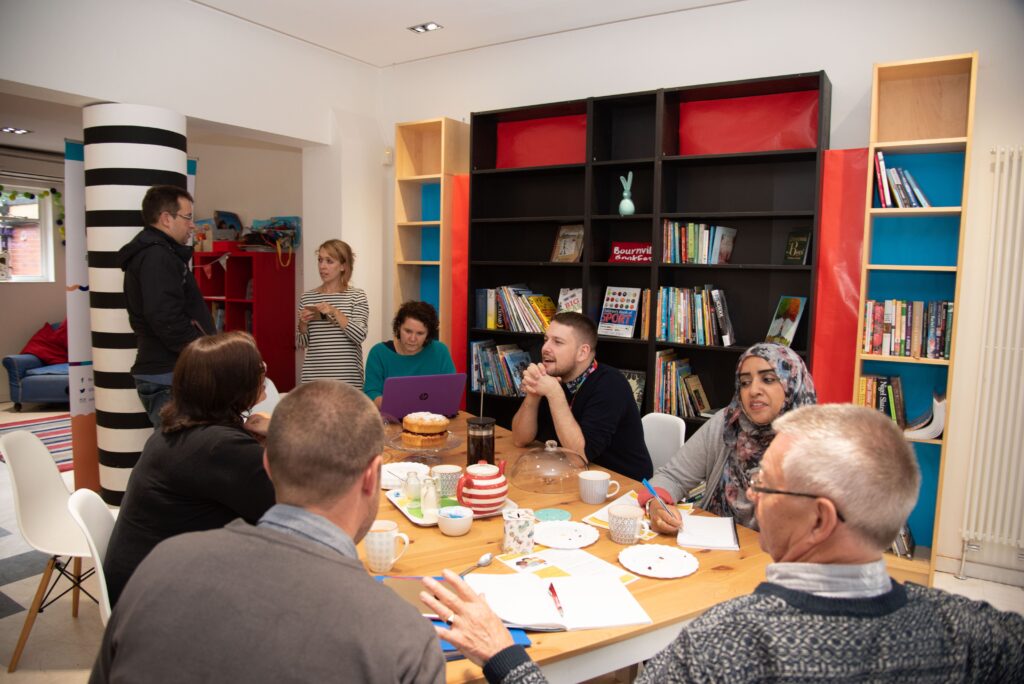 Eight people talking around a table, in a light and airy bookshop setting. Teapots and plates on the table, with cake crumbs